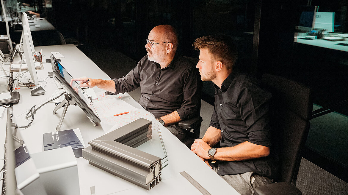  Zwei Männer in schwarzer Kleidung arbeiten gemeinsam an einem Computerarbeitsplatz.